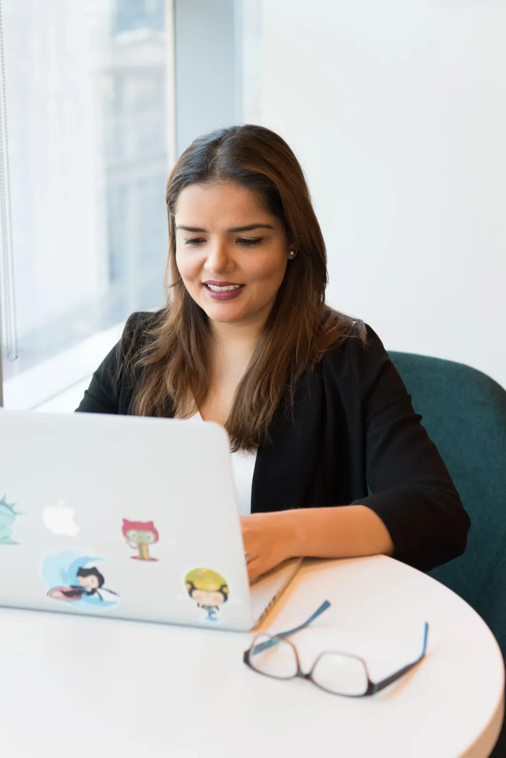 A smiling woman working on her laptop beside a window